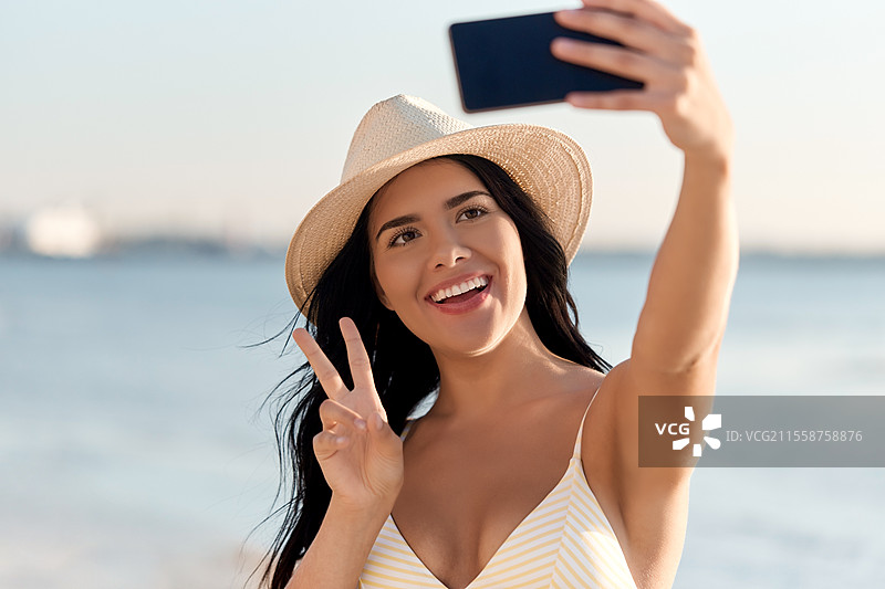 夏天和泳装概念 - 开心微笑的年轻女性穿着比基尼和草帽在海滩上用智能手机自拍。穿着比基尼的微笑女性在海滩上自拍。图片素材