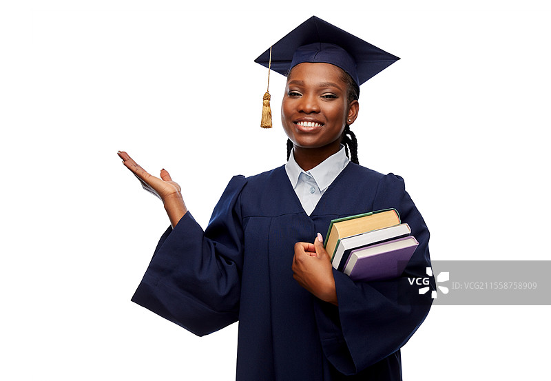 教育、毕业和人们的概念 - 开心的女毕业生穿着学士服和学位帽，手持书籍，向白色背景展示一些想象中的事物。开心的女毕业生手持书籍。图片素材