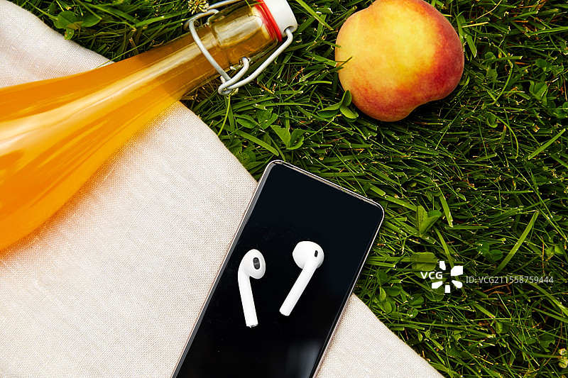 休闲、科技与夏季概念 - 近距离拍摄的智能手机与耳机、果汁瓶和桃子放在野餐毯上。智能手机和耳机放在野餐毯上。图片素材