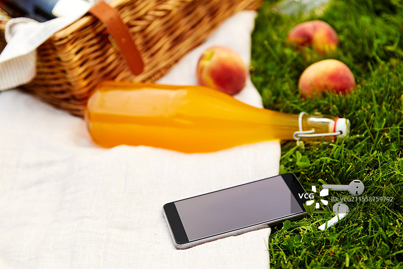 休闲、科技与夏季概念 - 草地上的野餐篮、果汁瓶和带耳机的智能手机特写。手机、耳机、野餐篮和果汁瓶。图片素材