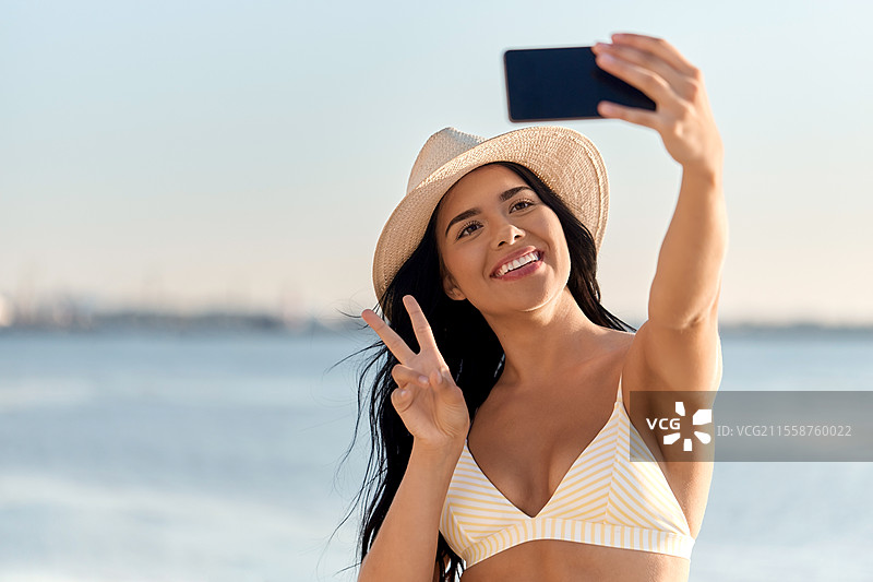 夏季和泳装概念 - 快乐微笑的年轻女性穿着比基尼泳衣和草帽在海滩上用智能手机自拍。穿着比基尼的女性在海滩上自拍。图片素材