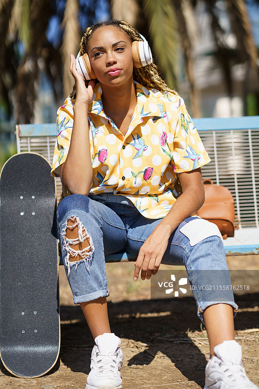 穿着休闲服的黑人女性坐在长椅上，旁边有滑板，戴着无线耳机听音乐，享受阳光明媚的夏日公园时光。非裔美国女性滑板者在听音乐。图片素材