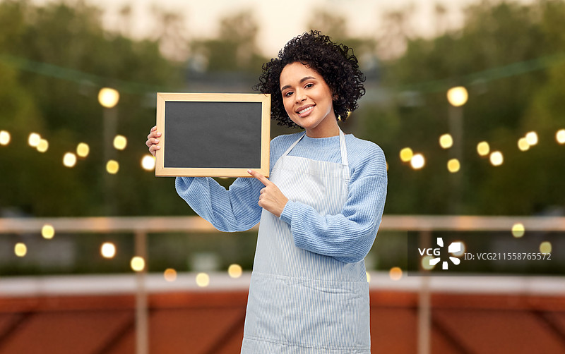 工作、职业和人们的概念 - 开心微笑的女性穿着围裙，手持黑色黑板，背景是餐厅露台或屋顶。开心的女性在餐厅露台上拿着黑板。图片素材