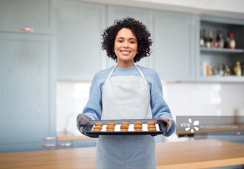 烹饪、烹饪艺术和人们的概念 - 开心微笑的女性穿着围裙，手里拿着装有燕麦饼干的烤盘，背景是家庭厨房。女性在厨房里拿着装有饼干的烤盘。图片素材