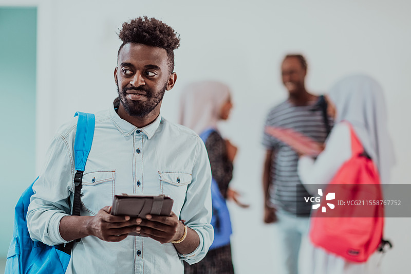 大学生活中，帅气的年轻学生手持平板电脑，微笑着站在大学前，身后与朋友们进行团队会议。高质量照片。图片素材