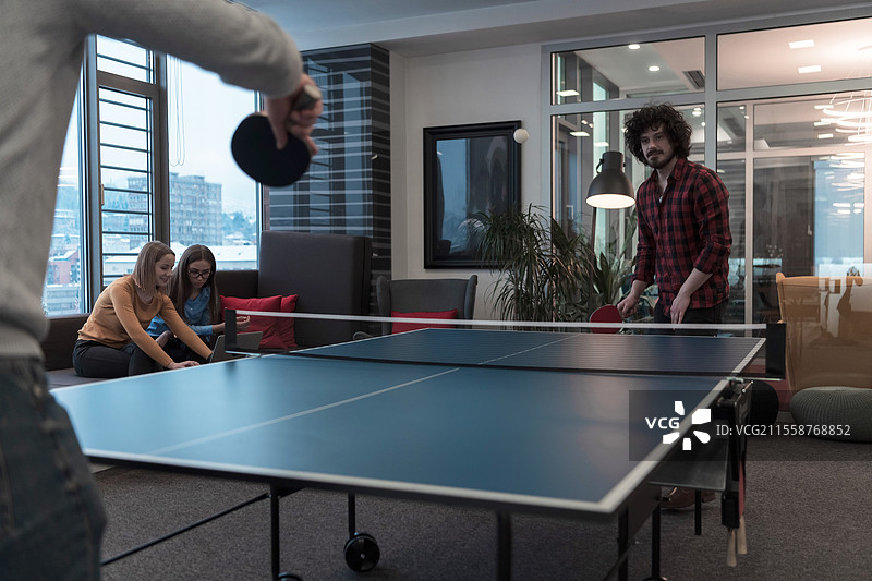 两位年轻的创业商人在现代创意办公室空间打乒乓球，背景中有一群人在开会和头脑风暴。高质量照片。图片素材