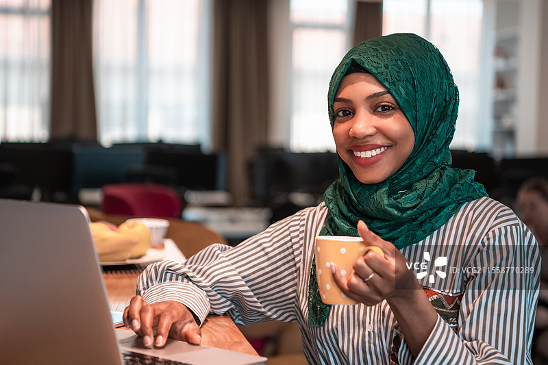 身穿绿色头巾的非洲穆斯林女商人在现代开放式创业办公室的休息区喝茶，同时使用笔记本电脑工作。高质量照片。图片素材