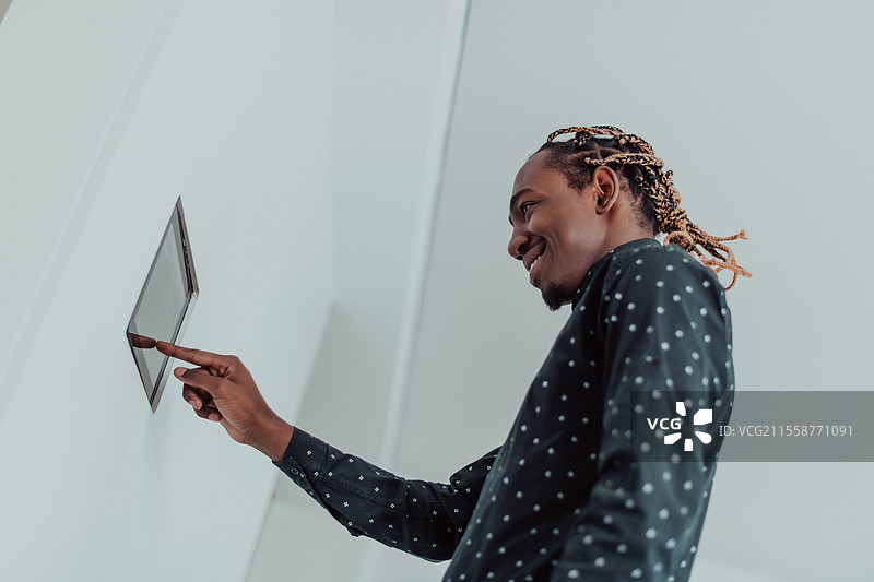 微笑的非洲裔美国男子使用现代智能家居系统，墙上的控制器，积极的年轻人调节恒温器的温度或在公寓中激活安全警报。高质量照片。图片素材