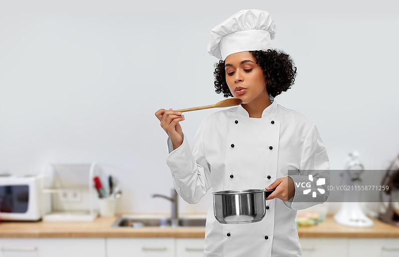 烹饪与人文概念 - 戴着厨师帽的女性厨师在厨房背景下用锅和勺子烹饪并品尝食物。女性厨师用锅和勺子烹饪食物。图片素材