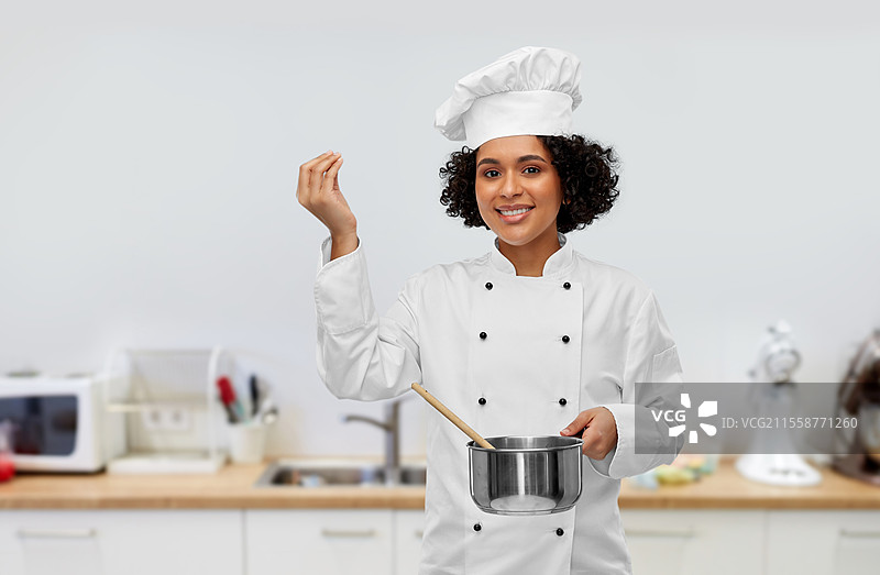 烹饪与人文概念 - 开心微笑的女厨师戴着厨师帽，手持平底锅在厨房背景下烹饪食物，展示美食标志。女厨师用平底锅和勺子烹饪食物。图片素材