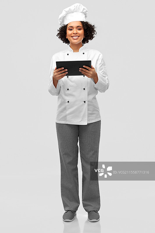 烹饪、烹饪艺术和人们的概念 - 开心微笑的女性厨师穿着厨师帽和外套，手持平板电脑，背景为灰色。微笑的女性厨师与平板电脑。图片素材