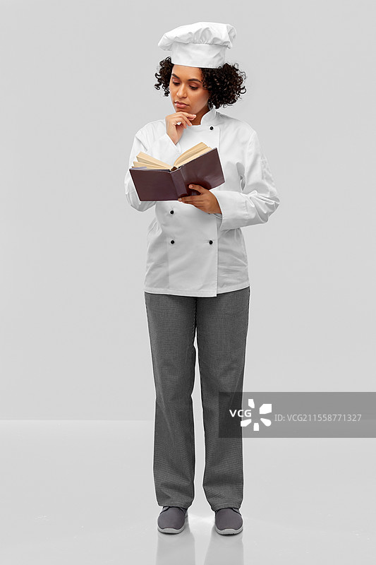 烹饪、烹饪艺术和人们的概念 - 一位穿着白色厨师帽和外套的女性厨师在灰色背景下思考，正在阅读食谱。穿着白色厨师帽的女性厨师正在阅读食谱。图片素材