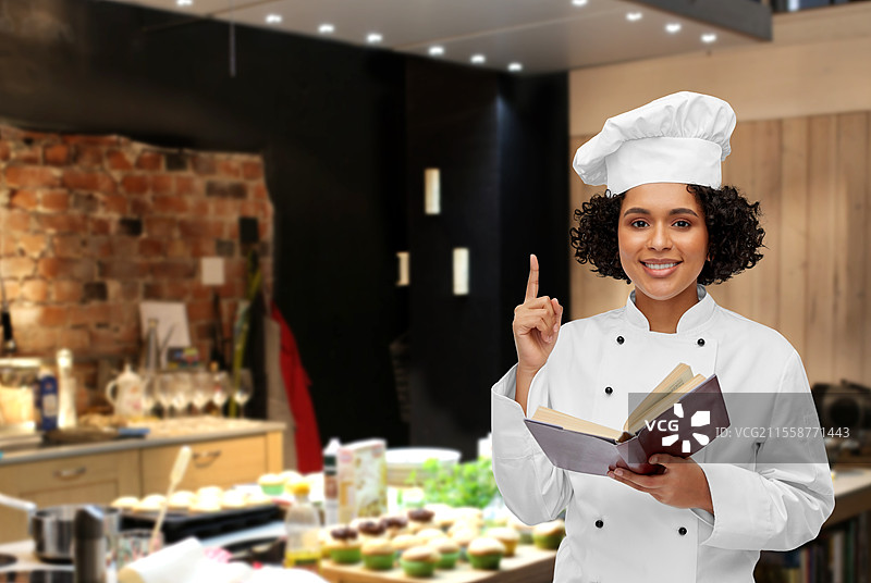 烹饪、烹饪艺术和人们的概念 - 开心微笑的女性厨师穿着白色厨师帽和外套，在餐厅厨房背景下阅读食谱。开心的女性厨师手持食谱。图片素材