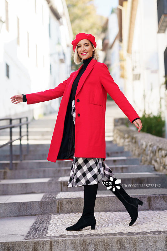 穿着时尚外套、连衣裙和贝雷帽的快乐金发女性，穿着靴子在街道台阶上走着，面带微笑地看着镜头。时尚装扮的女士在街道台阶上。图片素材