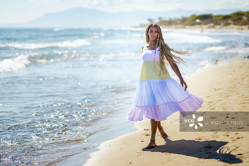 全身赤脚的非裔美国女性穿着夏季连衣裙，长发编成辫子，在阳光明媚的度假村海滩上走在沙滩上，海浪轻轻拍打着岸边。图片素材