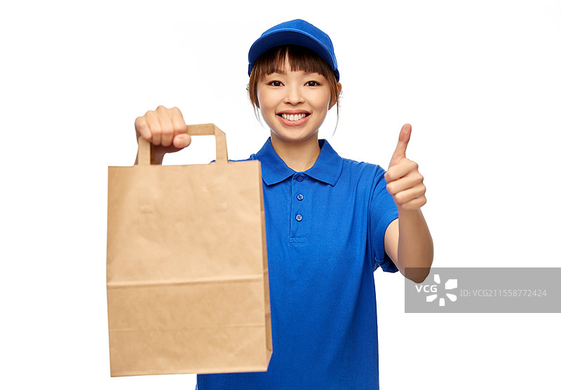 服务与工作概念 - 穿着蓝色制服、面带微笑的送餐员手持纸袋外卖，竖起大拇指，背景为白色。送餐员手持纸袋竖起大拇指。图片素材