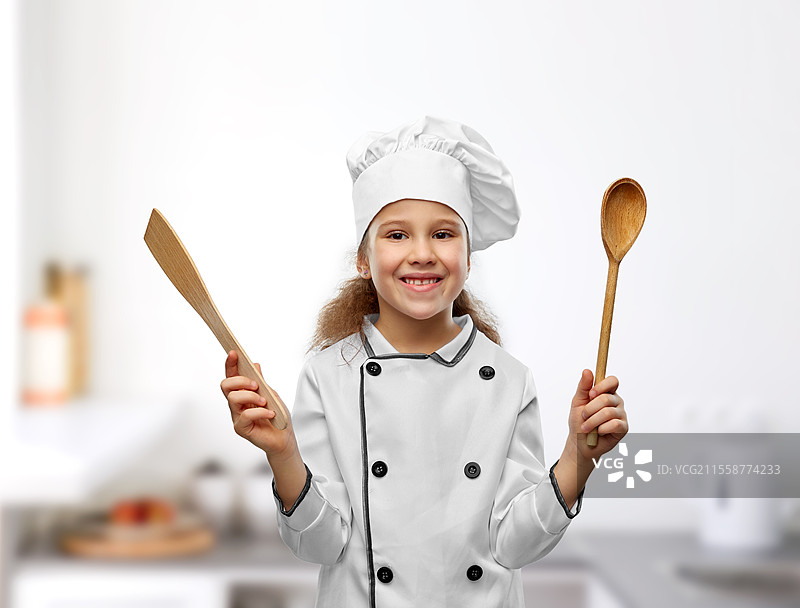 烹饪、烹饪艺术和职业概念 - 开心微笑的小女孩穿着厨师帽和外套，手持木勺和铲子，背景是厨房。开心的小女孩戴着厨师帽，手握勺子和铲子。图片素材
