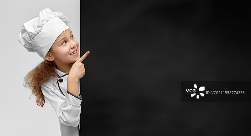 烹饪、烹饪艺术和职业概念 - 开心微笑的小女孩穿着厨师帽和外套，站在灰色背景的大黑板前。开心的小女孩戴着厨师帽，旁边有黑板。图片素材