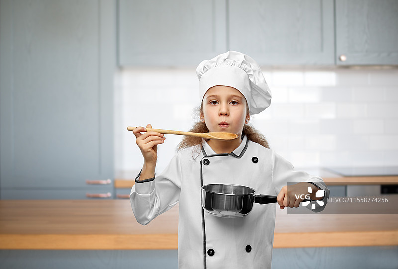 烹饪、美食和职业概念 - 小女孩穿着厨师帽和外套，手持平底锅，吹着木勺，背景是厨房。女孩戴着厨师帽，手握勺子和平底锅。图片素材