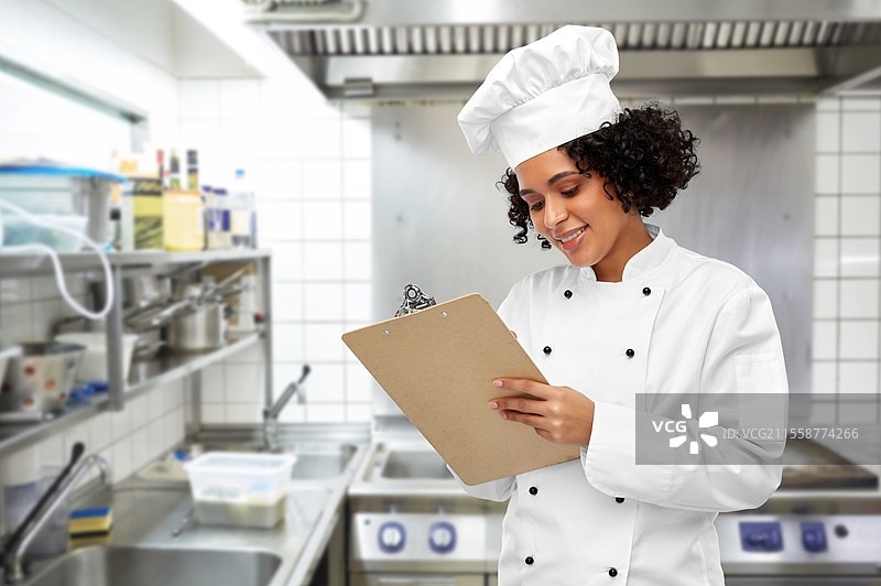 烹饪、烹饪艺术和人们的概念 - 开心微笑的女厨师穿着厨师帽和外套，手持夹板，背景是餐厅厨房。微笑的女厨师手持夹板。图片素材