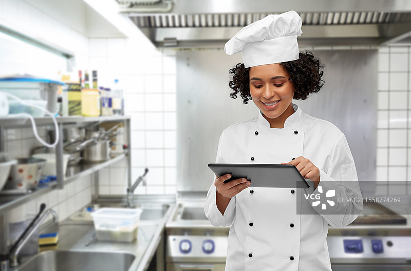 烹饪、烹饪艺术和人们的概念 - 开心微笑的女厨师穿着厨师帽和外套，手持平板电脑，背景是餐厅厨房。微笑的女厨师与平板电脑。图片素材