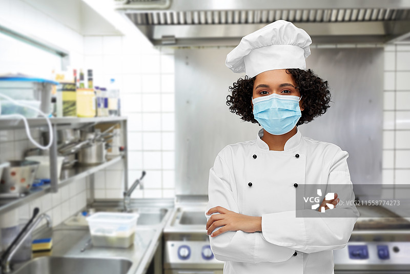 烹饪、疫情与健康概念 - 开心微笑的女厨师在餐厅厨房背景下佩戴防护口罩。女厨师戴着医用口罩和厨师帽。图片素材