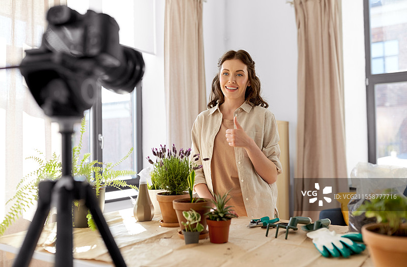 博客、园艺和家务概念 - 快乐的女性或博主手持相机和花盆录制教程视频，并在家中竖起大拇指。快乐的女性或博主在家中种植花卉。图片素材
