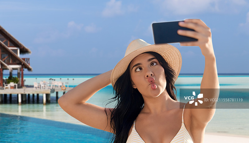 旅行、旅游和夏季假期概念 - 快乐微笑的年轻女性穿着比基尼泳衣和草帽，在法属波利尼西亚的热带海滩背景下用智能手机自拍。微笑的女性在海滩上自拍。图片素材