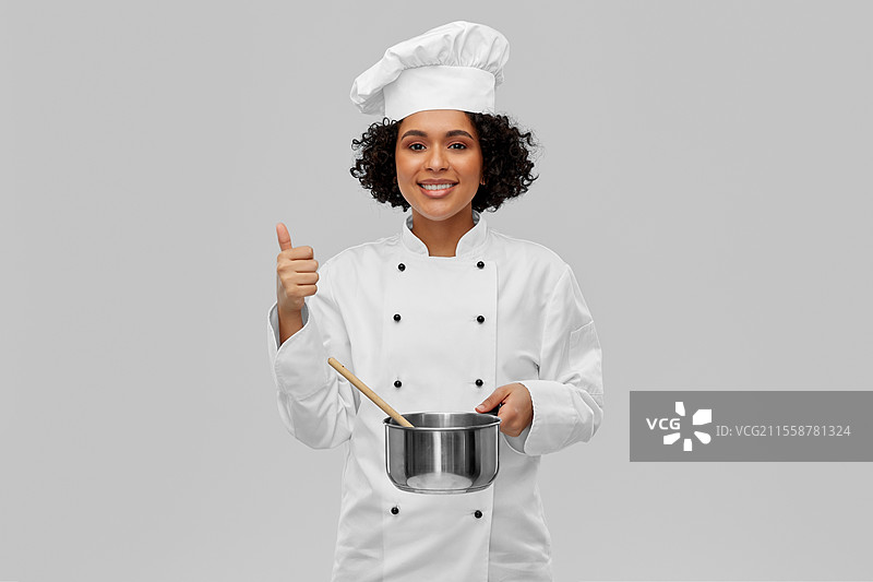烹饪与人们的概念 - 开心微笑的女厨师戴着厨师帽，手持平底锅在灰色背景下烹饪食物。女厨师用平底锅和勺子烹饪食物。图片素材