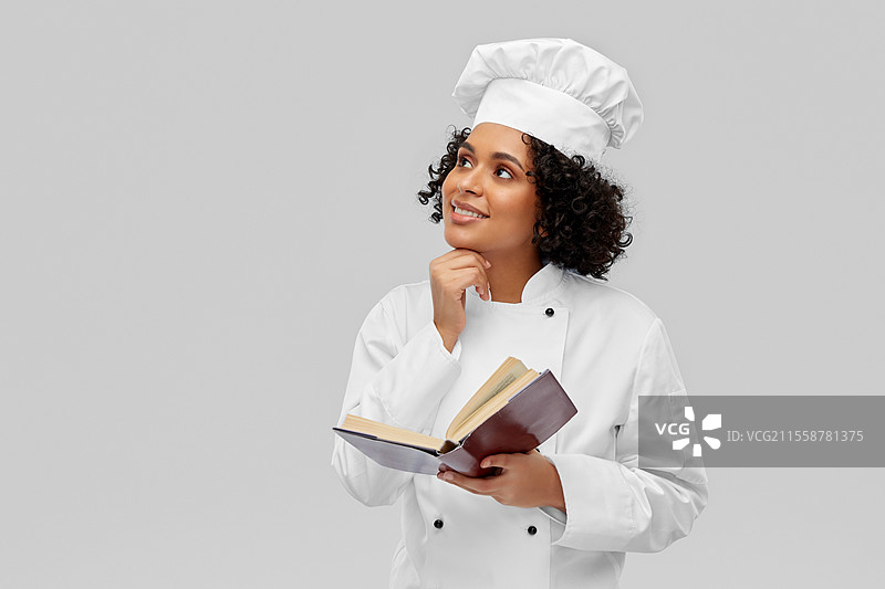 烹饪、烹饪艺术和人们的概念 - 开心微笑的女性厨师穿着白色厨师帽和外套，在灰色背景下阅读食谱。开心的女性厨师在阅读食谱。图片素材