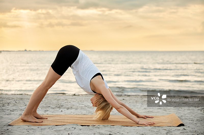 健身、运动和健康生活方式概念 - 女人在海滩上做日落时的瑜伽下犬式。图片素材