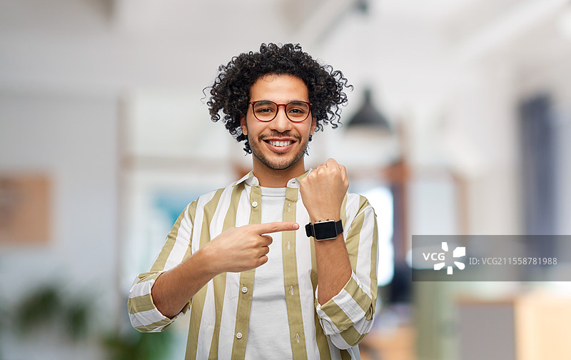 科技与人文概念 - 微笑的年轻男子戴着眼镜，在办公室背景下展示手腕上的智能手表。快乐的男子在办公室展示智能手表。图片素材
