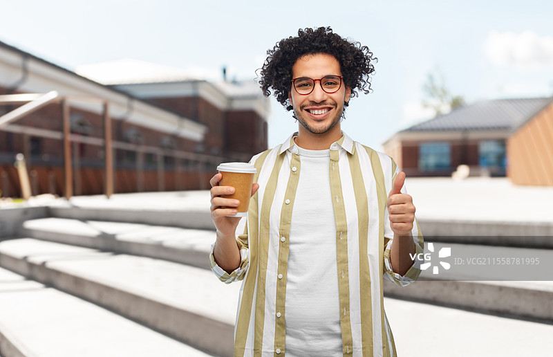 饮品与人群的概念 - 微笑的年轻男子戴着眼镜，手持外带咖啡杯，在城市街道背景下竖起大拇指。图片素材