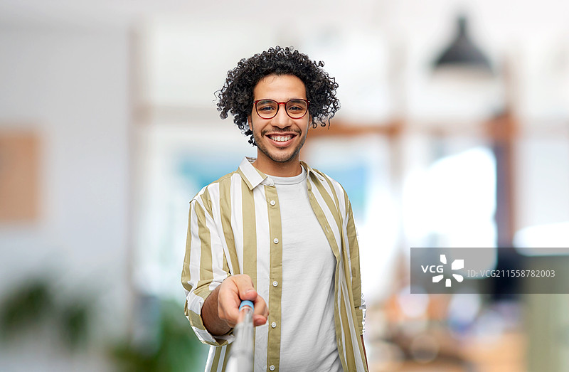 人们的概念 - 戴眼镜的年轻男子在办公室背景下开心地微笑，用自拍杆拍照。男子在办公室用自拍杆拍照。图片素材