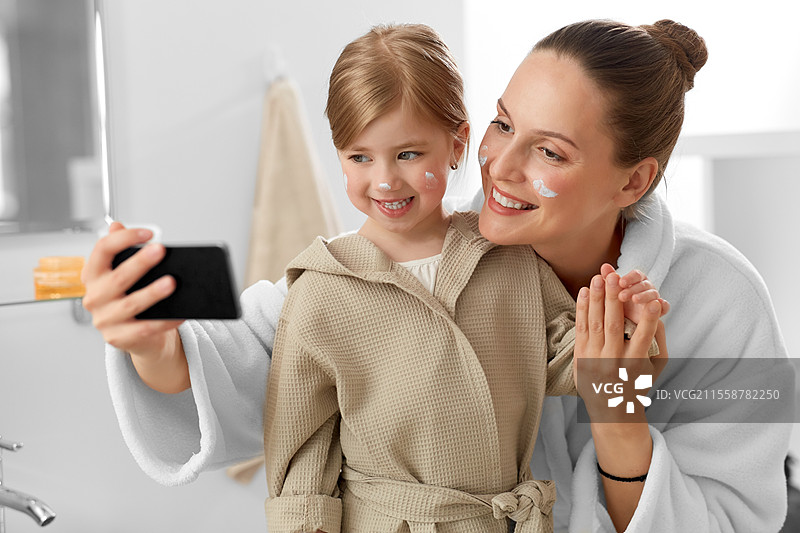 美容、家庭和人们的概念 - 开心微笑的母亲和女儿在浴室里用智能手机自拍，脸上涂抹着保湿霜。母女在浴室自拍。图片素材