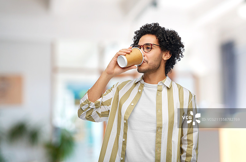 饮品与人群概念 - 戴眼镜的年轻男子在办公室背景下喝纸杯外带咖啡。男子在办公室喝外带咖啡。图片素材