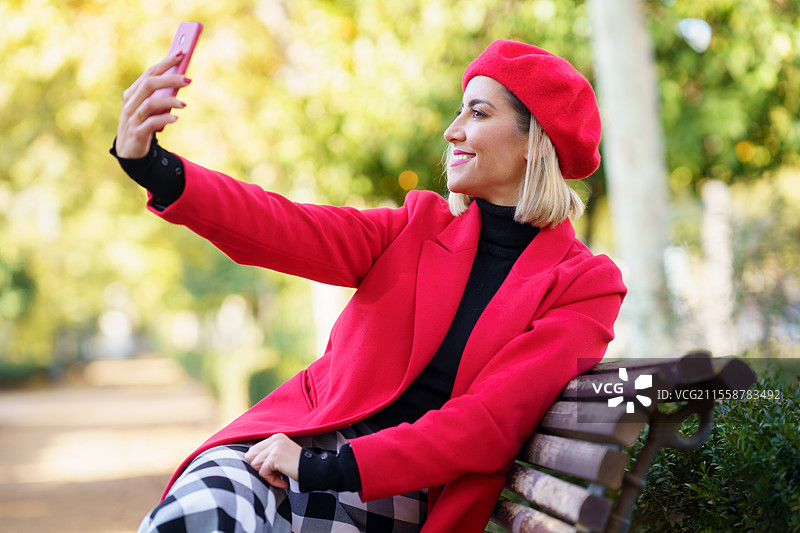 经典内容：金发女性在公园的木长椅上坐着，使用智能手机拍摄自拍，背景是模糊的树木。穿着红色衣服的快乐女性在公园里拍摄自拍。图片素材