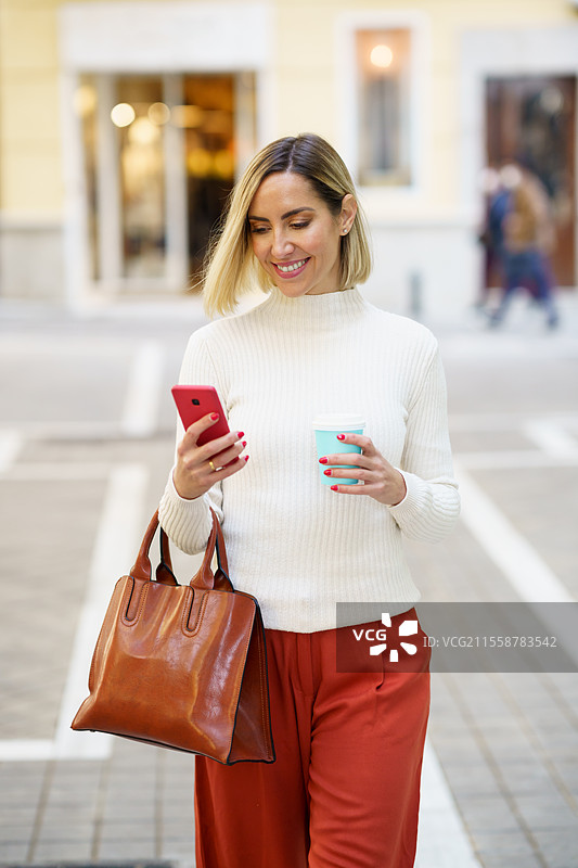 时尚打扮的快乐女性一边手拿饮料杯，一边在街上边走边浏览现代手机。积极的女性正在使用智能手机。图片素材