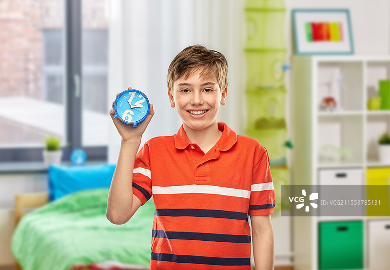 童年、时间管理和人际关系概念 - 穿着红色 polo 衫的快乐微笑男孩与闹钟的肖像，背景为家中的房间。家中快乐微笑的男孩与闹钟。图片素材