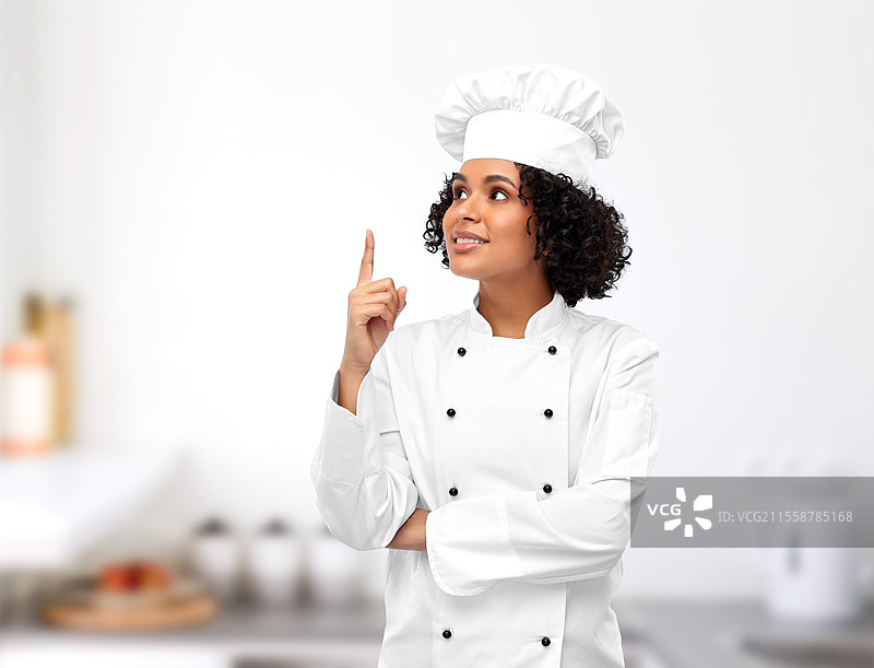 烹饪、广告和美食概念 - 开心微笑的女厨师戴着厨师帽，在餐厅厨房背景下指着上方。微笑的女厨师在厨房中指着上方。图片素材