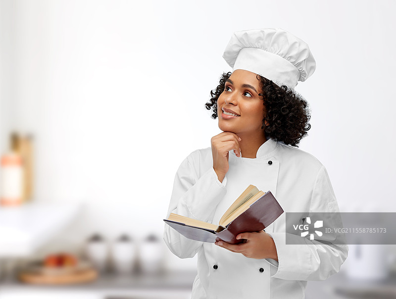 烹饪、烹饪艺术和人们的概念 - 开心微笑的女性厨师穿着白色厨师帽和外套，在餐厅厨房背景下阅读食谱。开心的女性厨师在厨房里翻阅食谱。图片素材