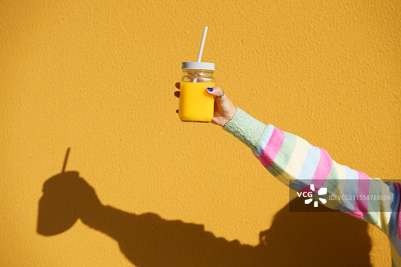 女性手握着一个带盖和吸管的玻璃杯，里面装满了新鲜的橙汁，背景为黄色。图片素材