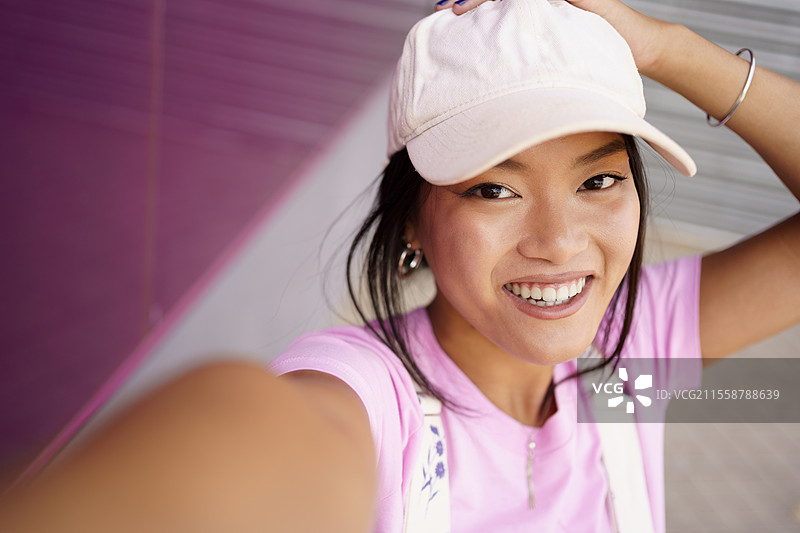 中国女孩用智能手机拍摄自拍照或视频，准备发布到社交媒体上。年轻的亚洲女性穿着现代服装和帽子。图片素材