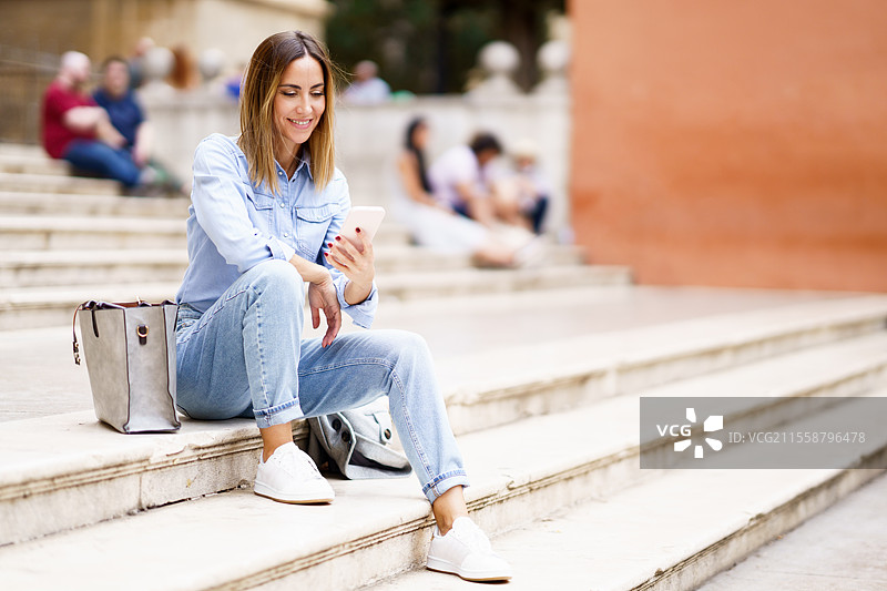 全身穿着休闲服的快乐女性，坐在城市街道的台阶上，微笑着用手机浏览社交媒体，旁边放着一个包。快乐的女性在台阶上使用智能手机。图片素材