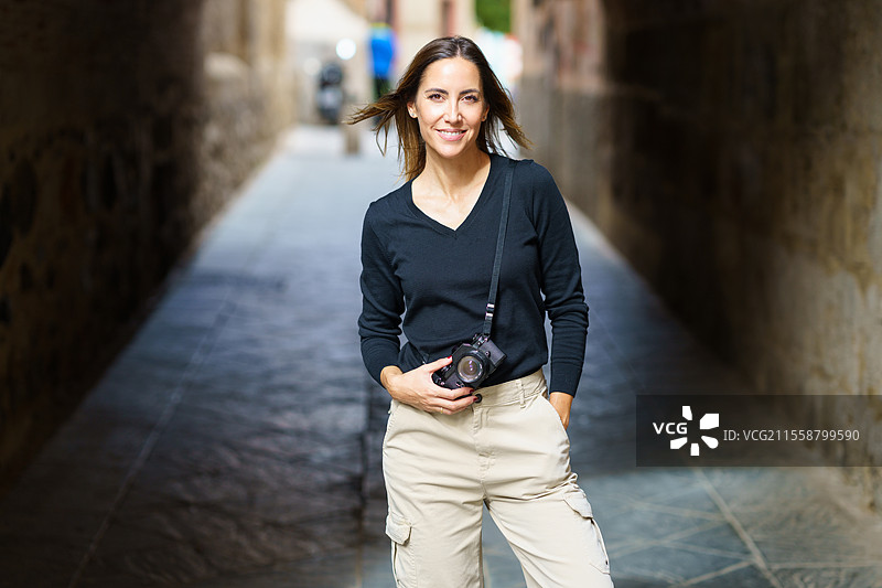自信的年轻女性摄影师穿着休闲服装，站在狭窄的街道上，手插在口袋里，微笑着看向镜头，享受假期。图片素材