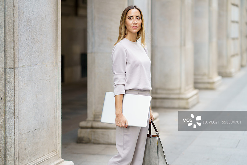 自信的成年女性，金发，穿着灰色服装，站在老建筑旁的街道上，手持笔记本电脑和手提包，目光投向远方。图片素材