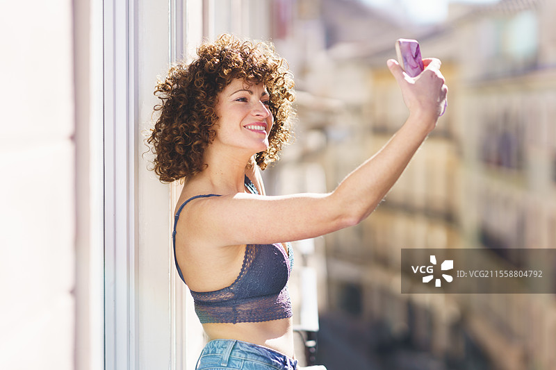阳光明媚的日子里，穿着文胸和牛仔裤的年轻女性在阳台上微笑着用智能手机自拍。图片素材