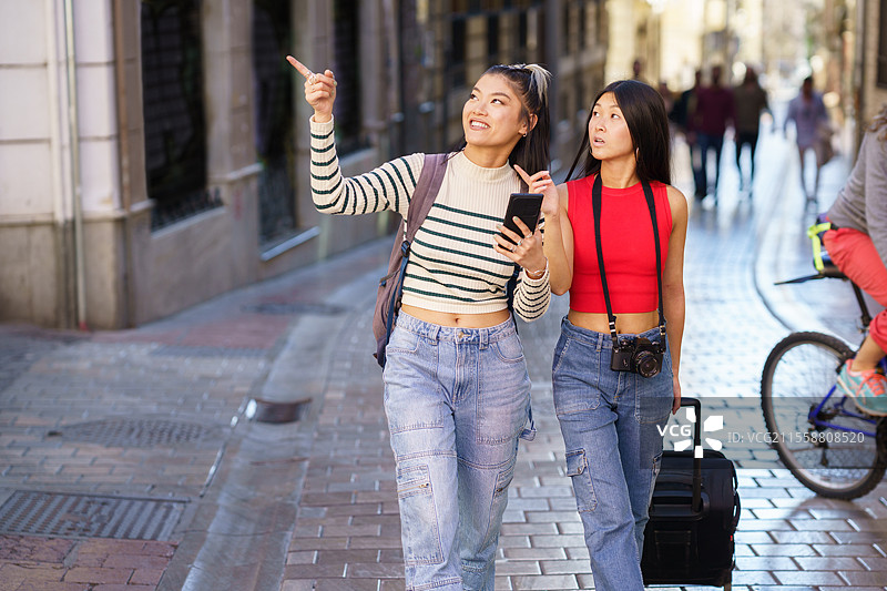 年轻积极的亚洲女孩背着背包和行李箱，在格拉纳达的街道上走着，一边用手机指着景点，享受旅程。亚洲女性旅行者在街上走着，指向远方。图片素材
