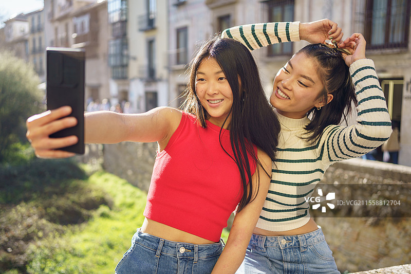 年轻的亚裔女孩们在格拉纳达的城市建筑前开心地微笑着，一起用手机拍自拍。快乐的亚洲女性朋友们在桥上一起拍摄自拍。图片素材