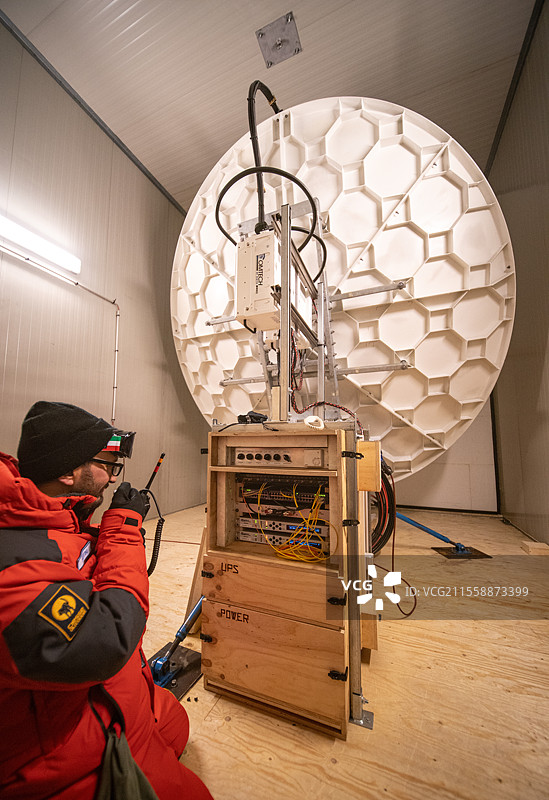 一名技术人员正在无线电上通话，蹲在一台电子设备控制台和一个大型无线电天线前。该技术人员与留在站内进行VSAT天线测试的搭档保持无线电联系，VSAT天线为站点提供互联网接入。电子设备被放置在一个定制的温控木柜中，以确保电子设备始终保持在0°C以上，以防止任何损坏。康科迪亚南极研究站，圆顶C高原，东南极。图片素材